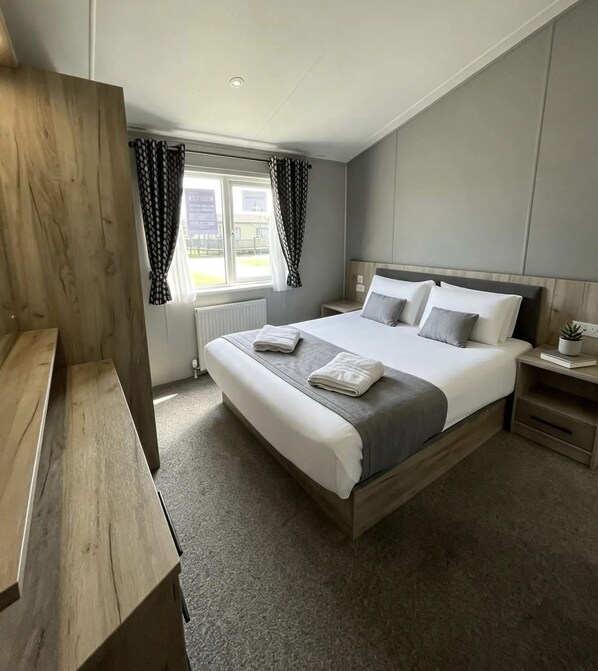 A modern bedroom with a large bed, a wooden bench, a bedside table with a book, and a window with curtains.