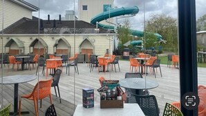 An outdoor patio area with tables and chairs, a water slide, and a building with arched windows in the background. - Sunset 5 -  Southview Holiday Park Skegness (Skegness)