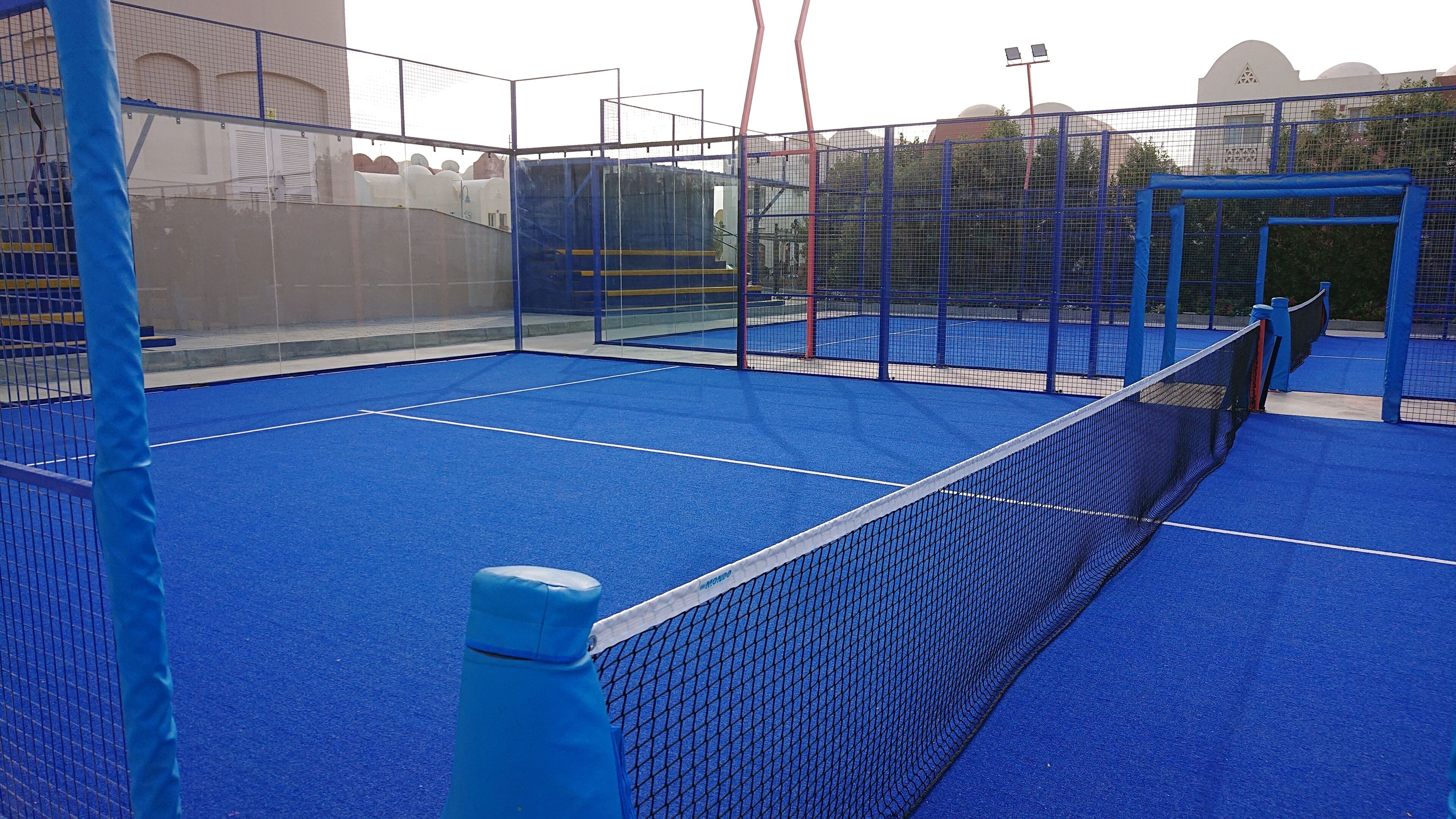 Un court de tennis intérieur doté d’un sol bleu et d’un filet, entouré de barrières bleues et agrémenté d’un espace de sièges abrité.