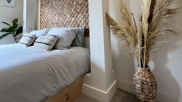 A bedroom with a bed, pillows, a woven vase with pampas grass, and a geometric wall panel.