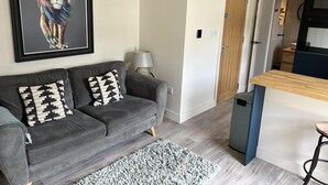 A living room with a grey sofa, patterned cushions, a framed artwork, a lamp, and a kitchen area with a wooden counter and bar stool.