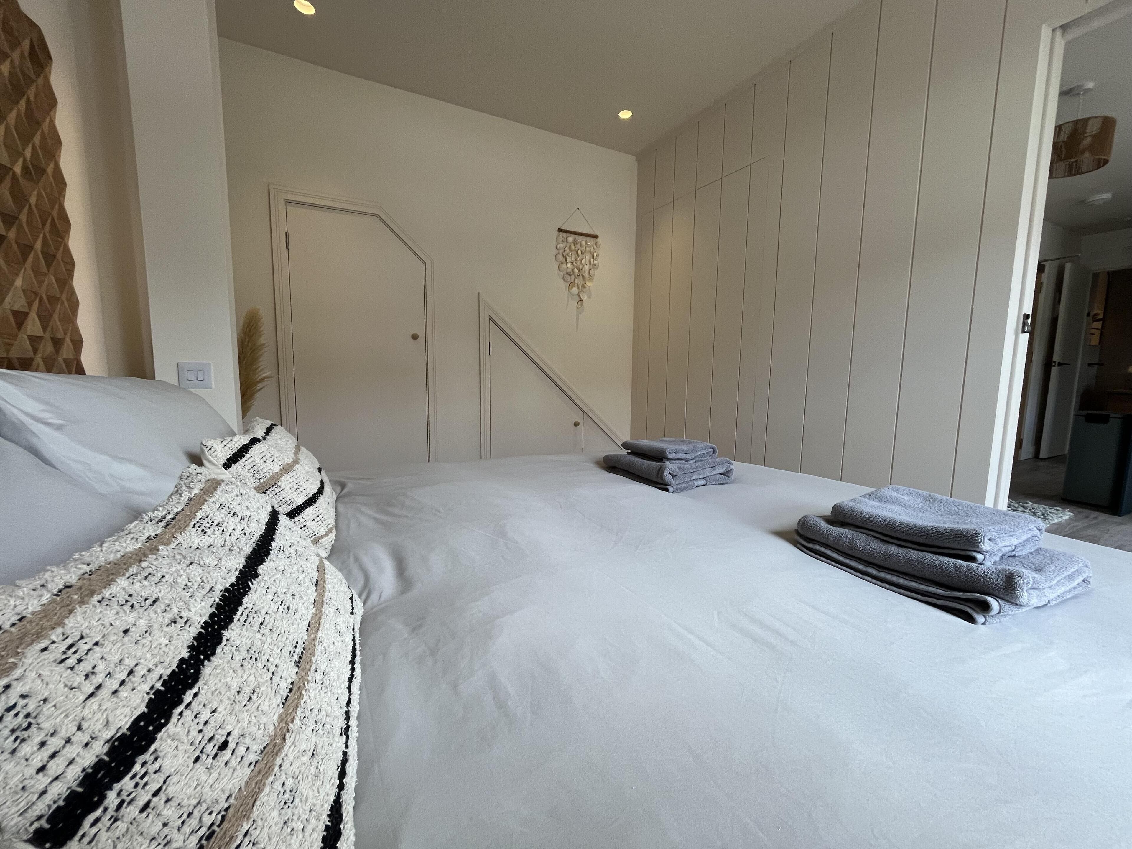 A neatly made bed with white linens and a crocheted blanket, a folded towel on the side, and a door leading to another room.