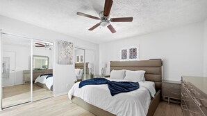 Quarto com cama, ventilador de teto, cômoda, espelho e janela.