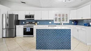 Uma cozinha moderna com armários brancos, geladeira de aço inoxidável, micro-ondas, forno e uma ilha revestida de azulejos azuis.