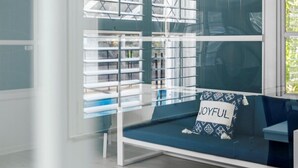 Quarto moderno com divisória de vidro, sofá azul com almofada 'JOYFUL' e vista para o exterior pela janela.