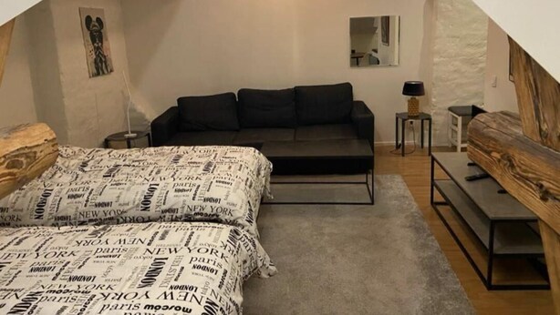 Een knus zolderkamer met een donkere bank, een bed met beddengoed met een patroon, een klein tafeltje en een spiegel aan de muur.