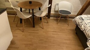 Een knus kamertje met een ronde houten tafel en vier witte stoelen. Een bed met een geborduurd kleed staat rechts. De vloer is van hout en de muren zijn wit met een gespikkelde afwerking.