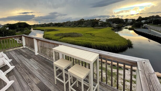 A wooden deck with a table and chairs overlooking a river and marshland with houses in the background.