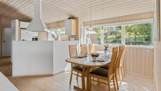 Une cuisine moderne avec des meubles en bois et un îlot central, un coin repas avec des chaises et une table en bois, et de grandes fenêtres offrant une vue sur la verdure.