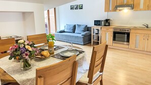 A modern kitchen with wooden cabinets and stainless steel appliances, a dining area with a table set for two, and a grey sofa in the background.