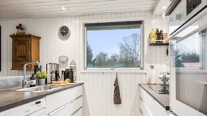 A modern kitchen with white cabinetry, a built-in dishwasher, a window with a view, and a wooden cabinet with a clock. - "Juste" - 1.7km from the sea by Interhome (Hals)