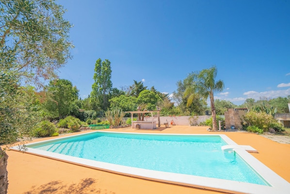 Relax by the serene pool surrounded by lush greenery and trees, with a shaded area beneath a wooden structure.