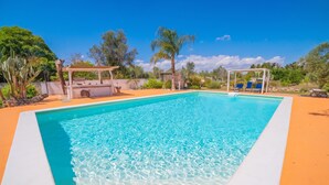 Una piscina rettangolare con acqua cristallina, circondata da una zona sabbiosa con un pergolato e poltrone da relax.