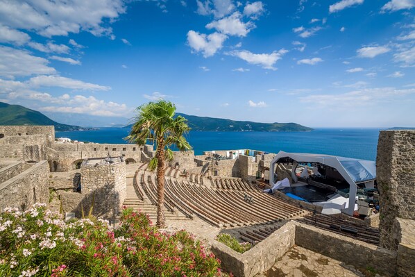 An ancient amphitheater with stone seating and a stage, overlooking a coastal area with a clear sky.