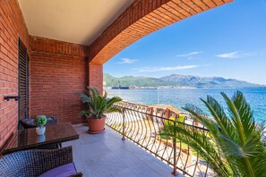 Ein Balkon mit Blick auf das Meer und die Berge, einem Tisch mit Stühlen und Topfpflanzen.