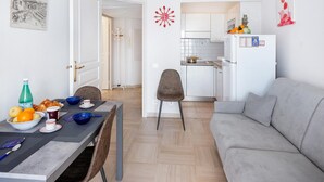 Ein modernes Wohnzimmer mit grauem Sofa, einem Essbereich mit für ein Frühstück gedecktem Tisch und einer Küche mit weißen Schränken und einem Kühlschrank.