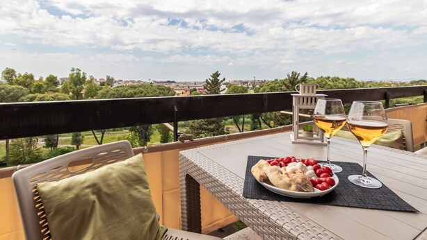 A balcony with a table set for a meal, featuring a plate of food and two glasses of wine, overlooking a cityscape.