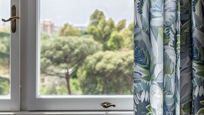 A window with a view of trees and buildings, a white door with a brass handle, and a curtain with a leaf pattern.