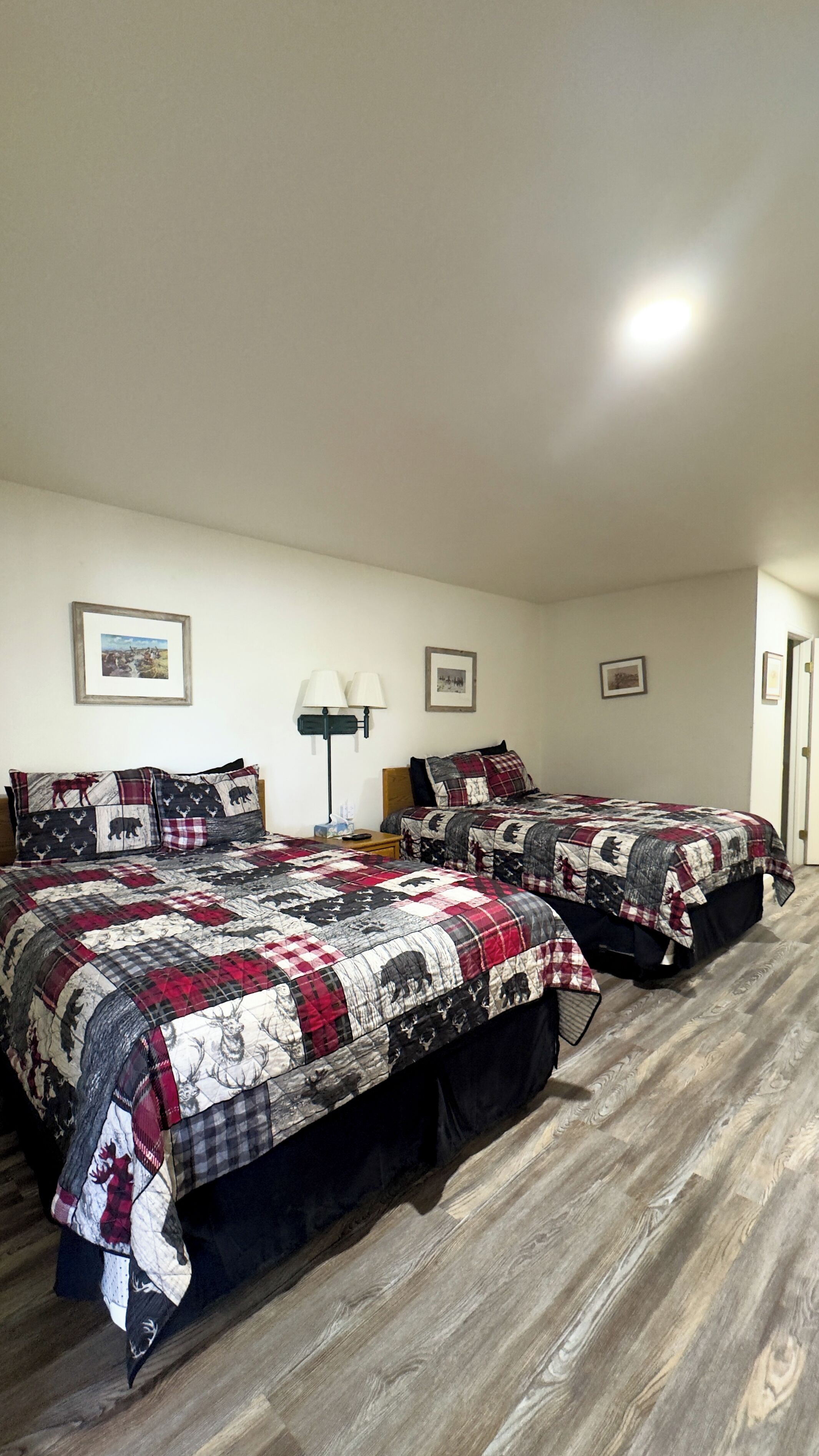 A room with two beds, each with a quilted bedspread featuring animal prints. There are framed pictures on the wall, a bedside lamp, and a door to the right.