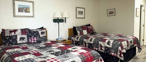 A room with two beds, each with a quilted bedspread featuring animal prints. There are framed pictures on the wall, a bedside lamp, and a door to the right.