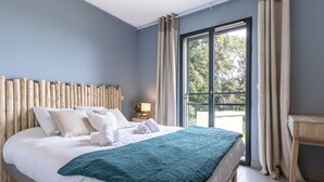 Ein modernes Schlafzimmer mit einem Holzkopfende, einem großen Bett mit weißen Leinen und einer türkisblauen Tagesdecke, einem Nachttisch mit einer Lampe und Blick auf Bäume durch ein Fenster mit Vorhängen.