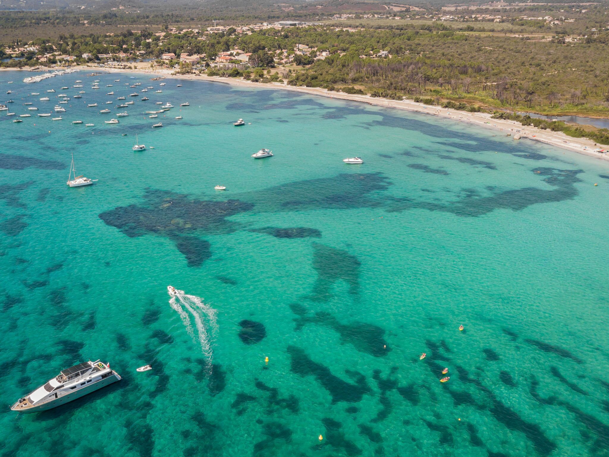 Vista aérea de una zona costera con aguas turquesas cristalinas, numerosos barcos y un yate grande anclado cerca de la orilla.
