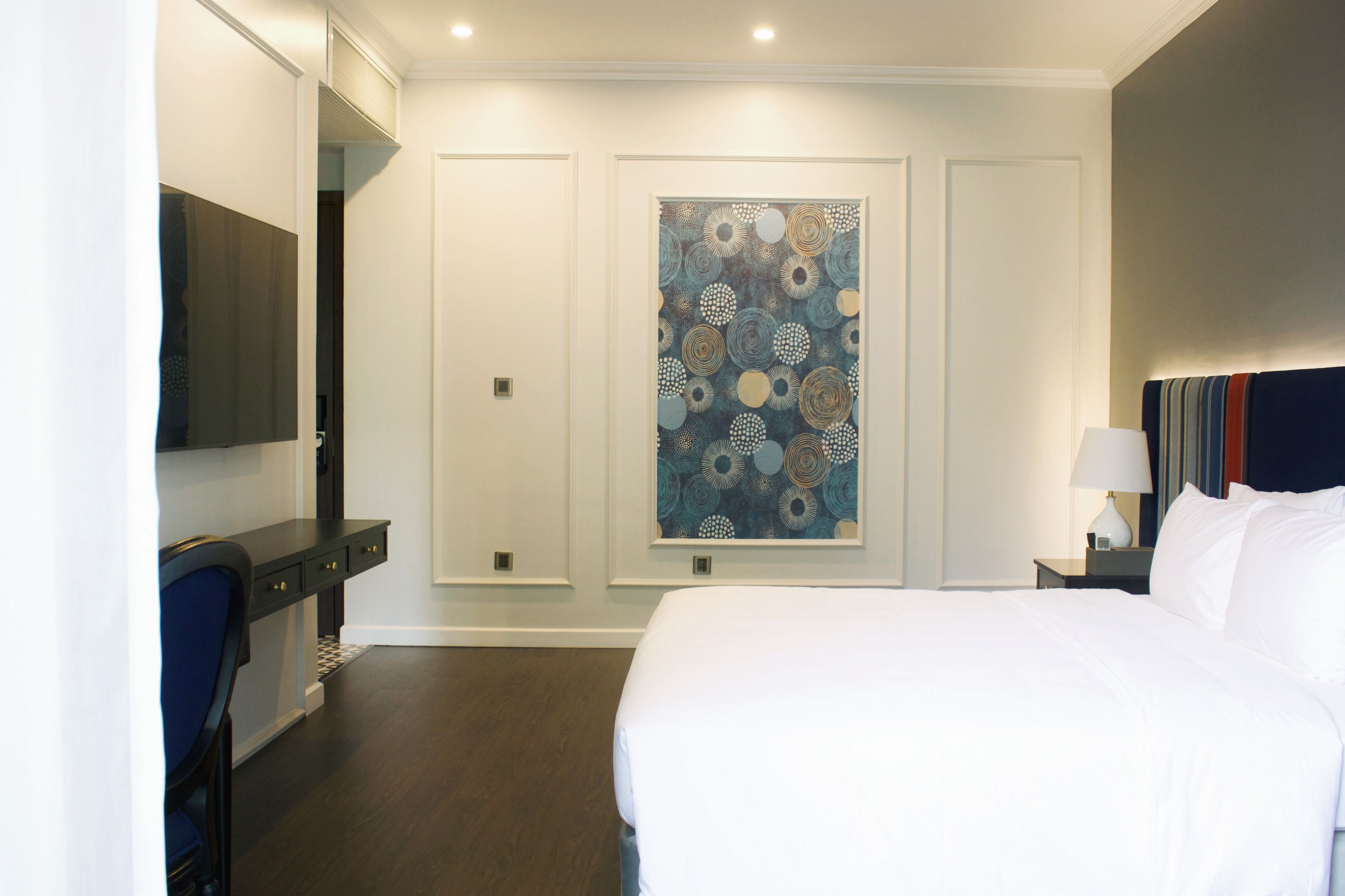 Une chambre d’hôtel comprenant un lit, un bureau, une chaise, un téléviseur fixé au mur et un grand tableau abstrait.