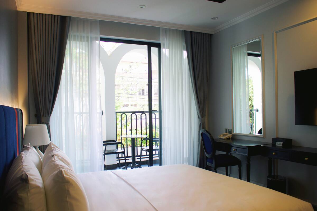 A hotel room with a bed, a desk, a chair, a lamp, and a balcony with a table and chairs.