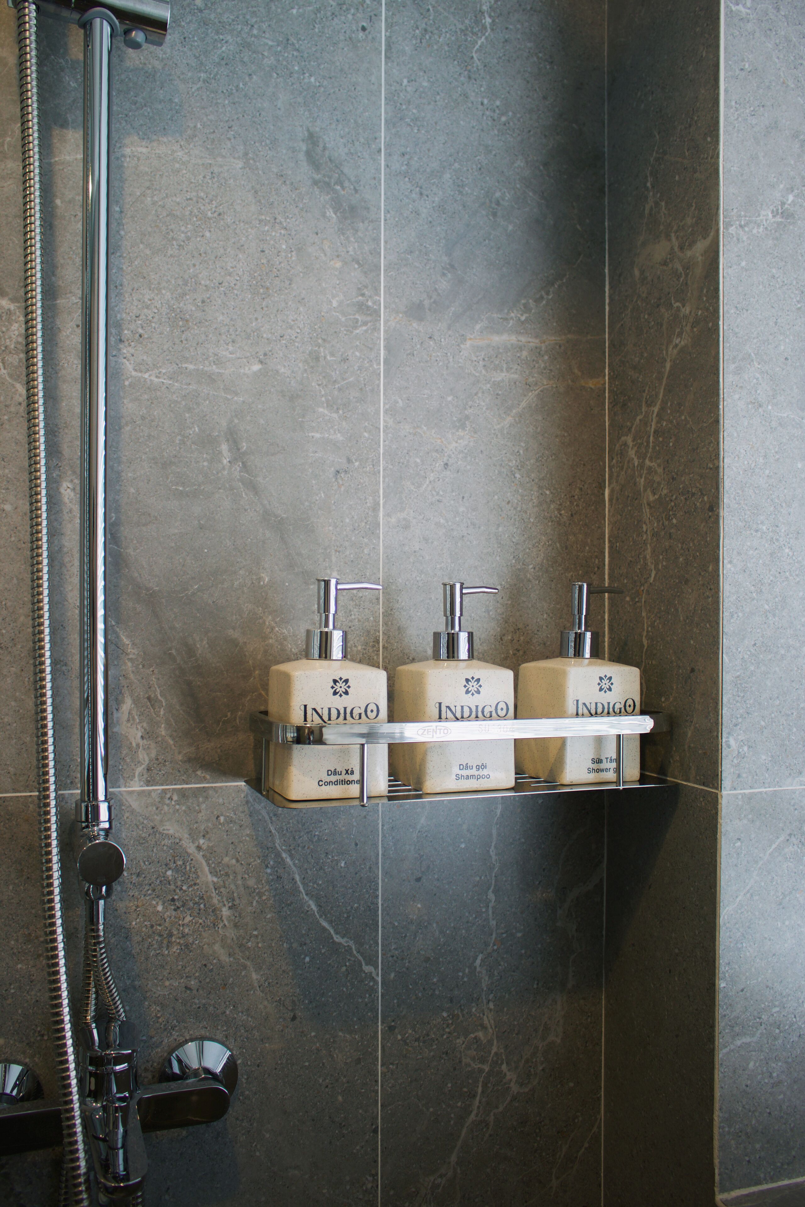 A bathroom with a shower, a hand shower, and three bottles of Indigo shampoo and conditioner on a shelf.