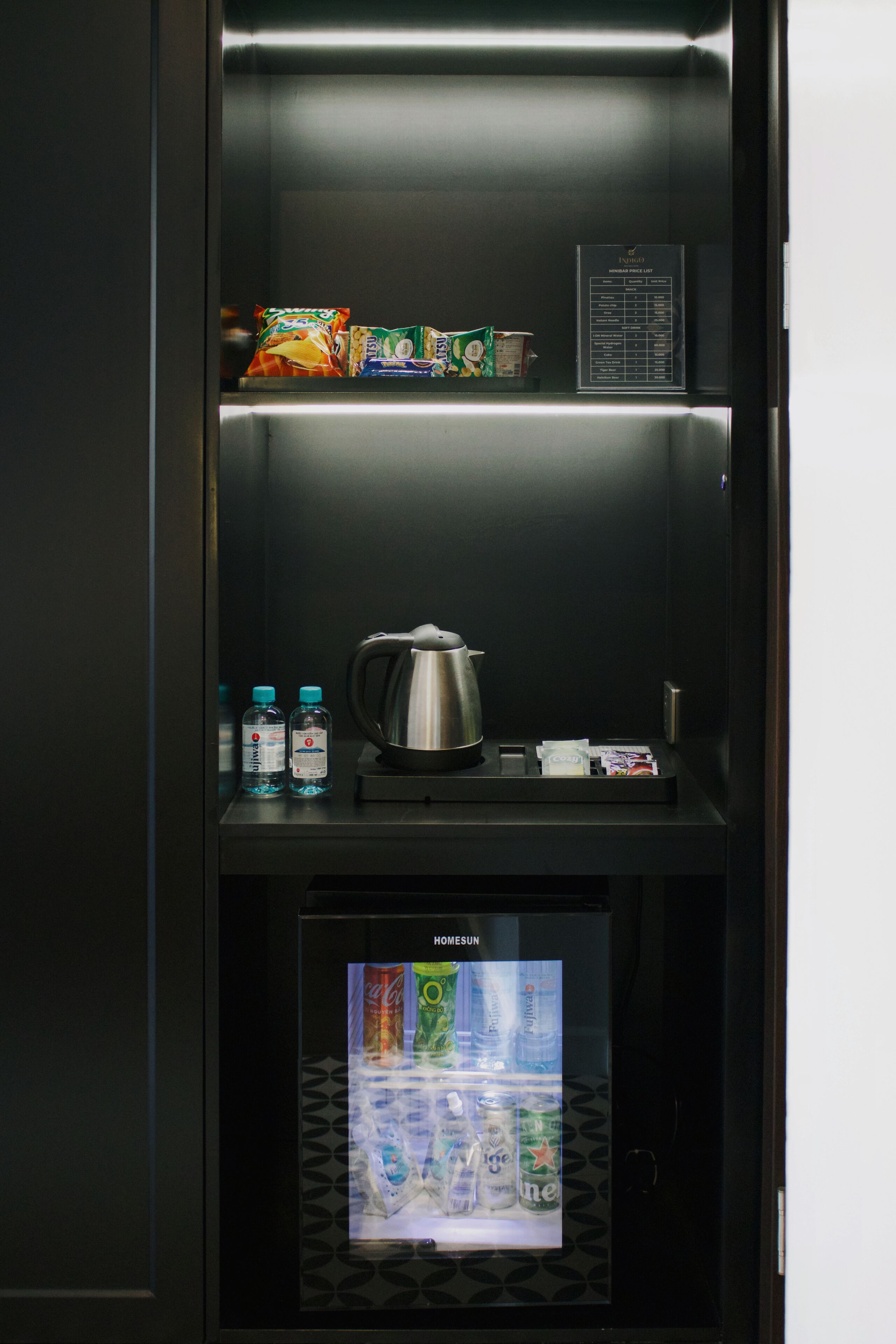 A minibar with snacks, bottled water, a kettle, and a refrigerator displaying beverages.