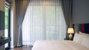 A hotel room with a large bed, a desk with drawers, a lamp on a bedside table, and sheer curtains on the window. - Indigen Boutique Hotel Cao Bang (Thuc Phan)