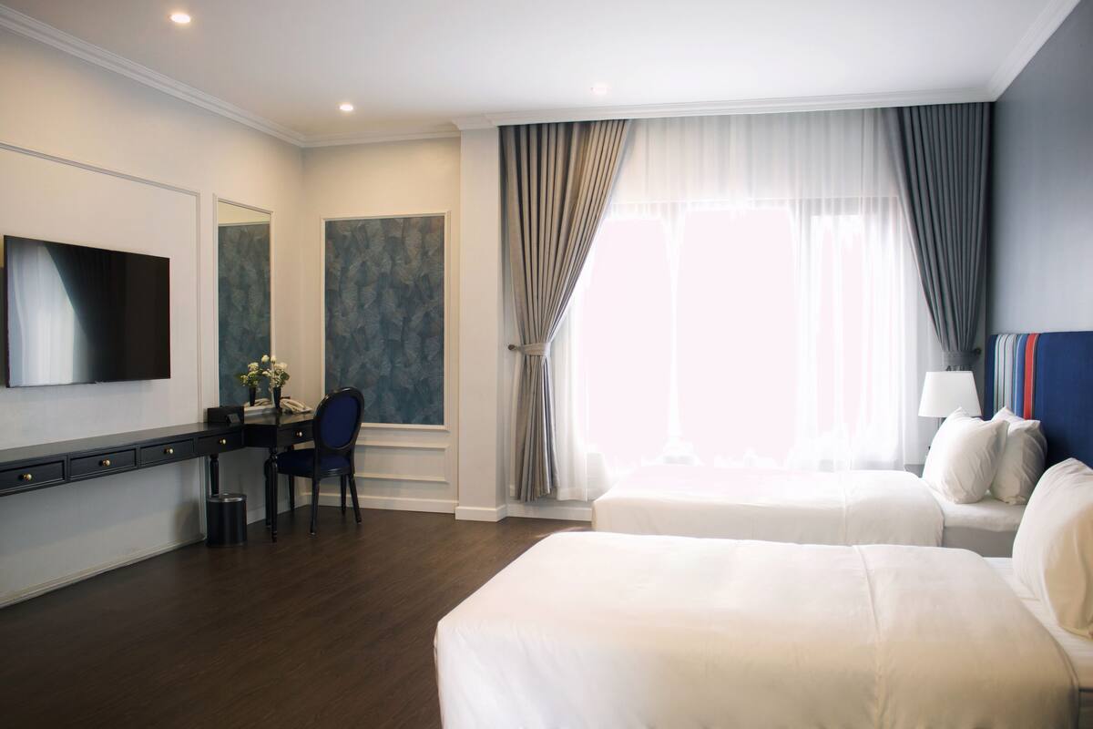 A hotel room with two beds, a desk with a chair, a TV mounted on the wall, and a large window with curtains.