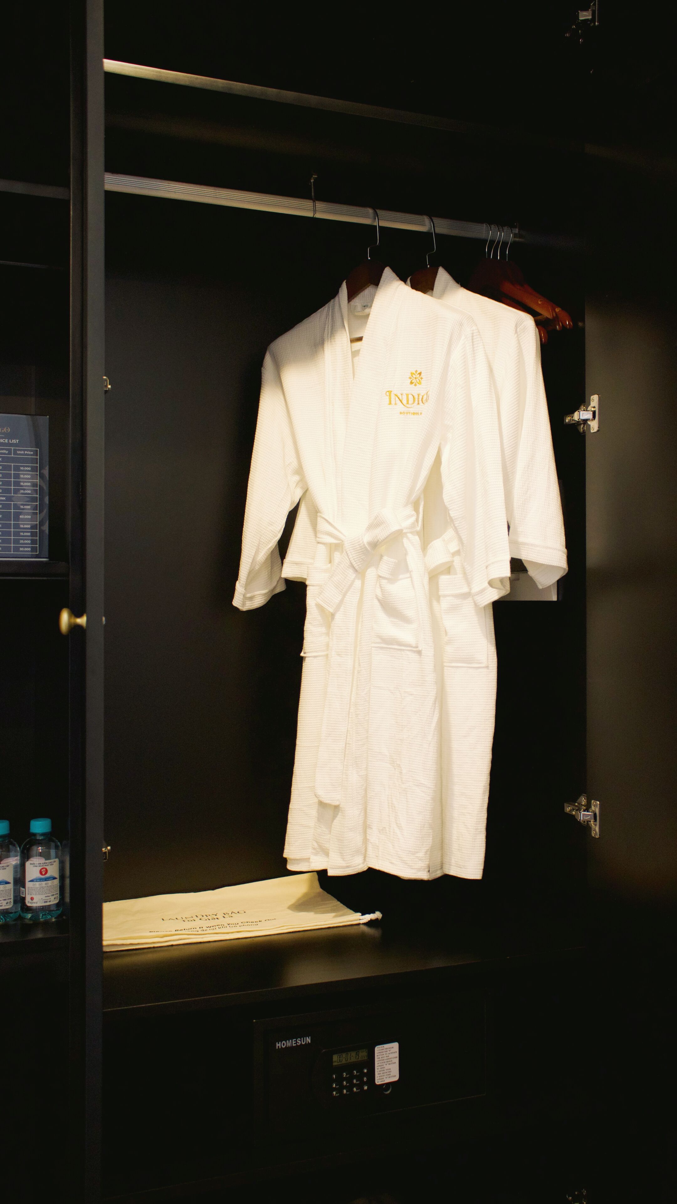 A white robe hanging in a dark room with a digital display and a bottle of water on the shelf.