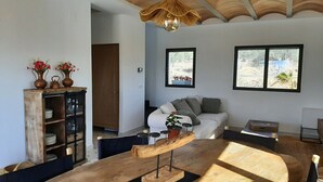 Ein modernes Wohnzimmer mit einem Holzesstisch, einer Couch und einer Holzkabine mit Glastüren. Zwei Fenster bieten Ausblicke nach draußen.
