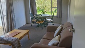 A living room with a brown sofa, a wooden coffee table, and a dining table with chairs. There is a window with curtains, an air conditioning unit, and a door. - 1970's Retro style 2 bed unit (Auckland)