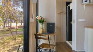 Una habitación con piso de madera, un escritorio con silla, una mesita con un florero y una puerta que da a otra habitación.
