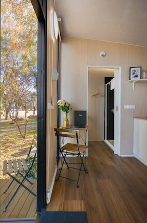 Une pièce avec un plancher en bois, un bureau avec une chaise, une petite table avec un vase et une porte qui donne sur une autre pièce.