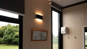 A room with a framed picture, a wall-mounted light, and a window with a view of greenery.