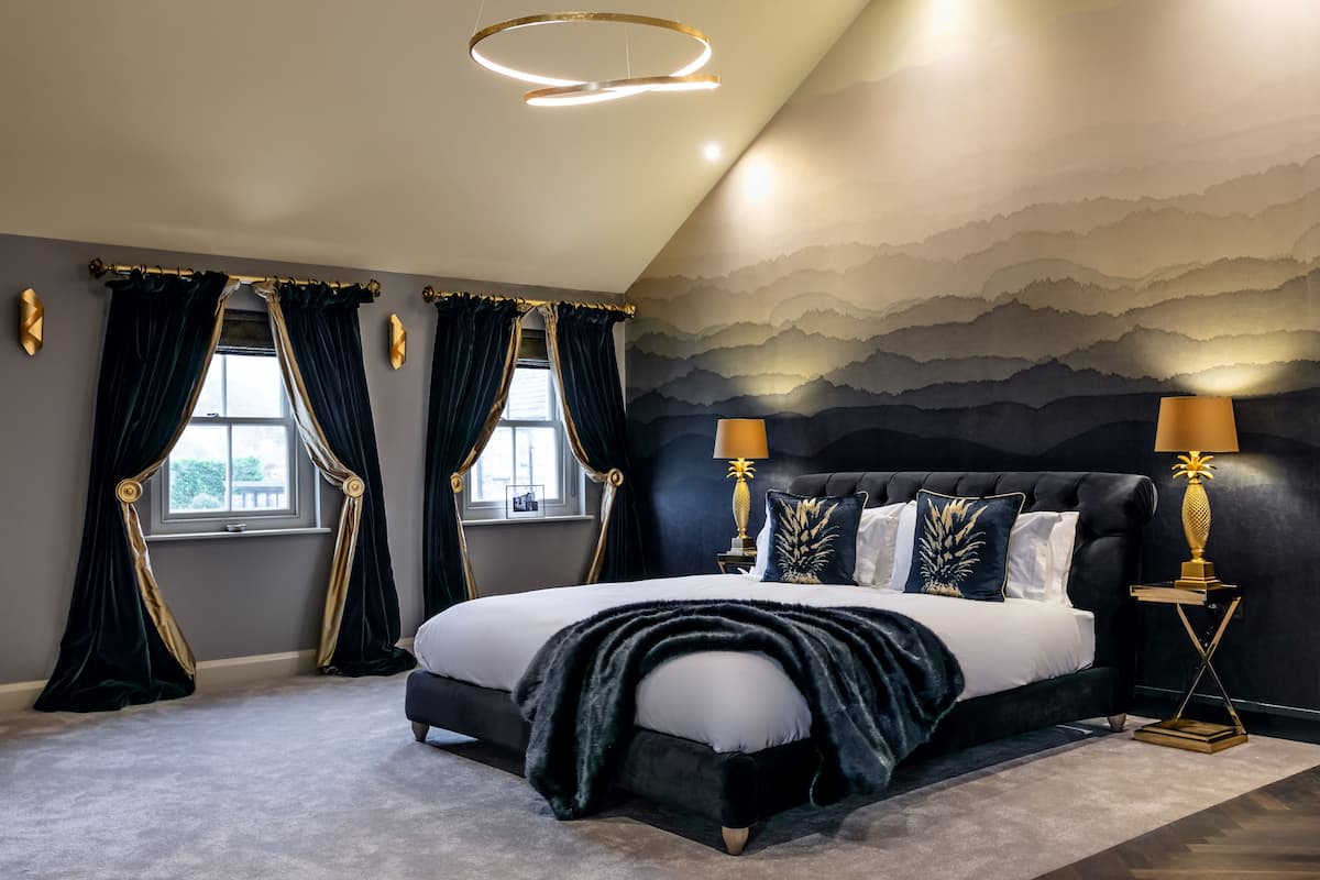 Quarto com cama grande, cortinas escuras, duas luminárias de cabeceira e papel de parede com tema de montanha.