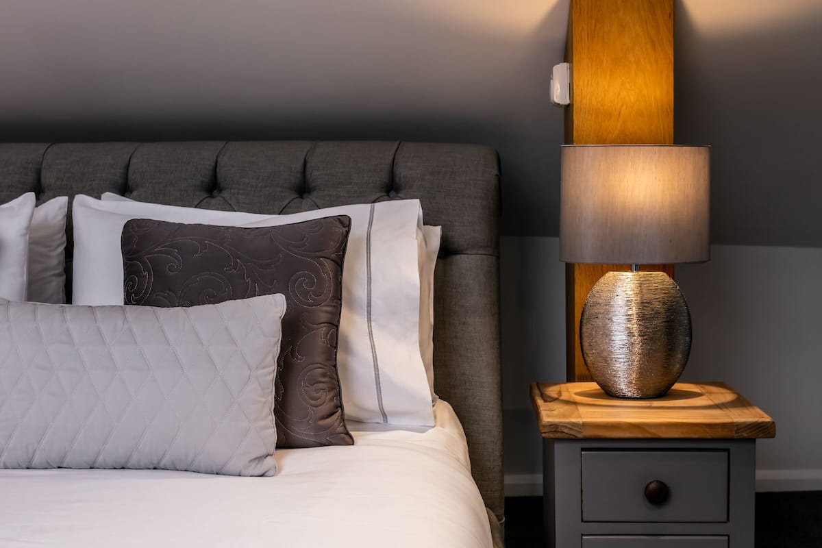 Uma cama bem arrumada com cabeceira estofada cinza, uma mesa de cabeceira de madeira com abajur e uma gaveta.