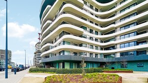A multi-story hotel with a modern design, featuring balconies on each floor, a landscaped garden area in the foreground, and a busy street with vehicles in the background.