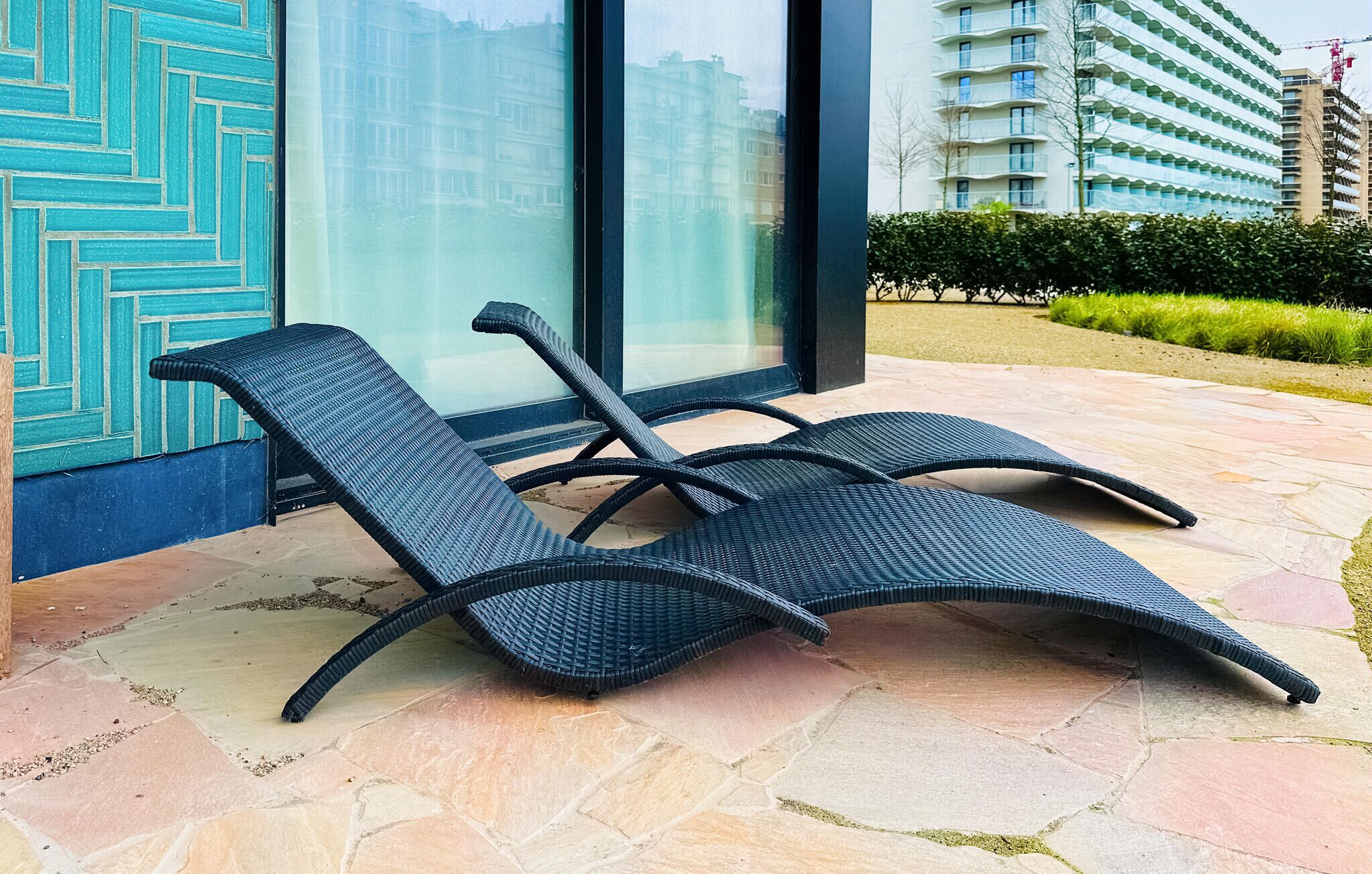 Un fauteuil de relaxation avec un siège et un dossier en osier, placé sur une terrasse en pierre offrant une vue sur un bâtiment moderne et un jardin aménagé.
