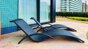 A lounge chair with a woven seat and backrest, positioned on a stone patio with a view of a modern building and a landscaped garden.