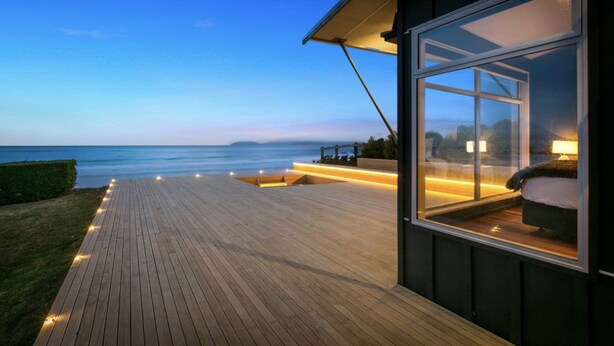 Una casa moderna con terraza de madera que da a un cuerpo de agua, un ventanal con una cama en su interior y un espacio exterior bien iluminado.