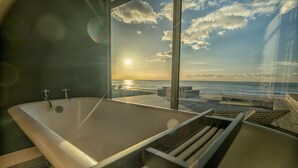 Un baño moderno con una bañera grande, cabina de ducha de vidrio y vistas al océano al atardecer.