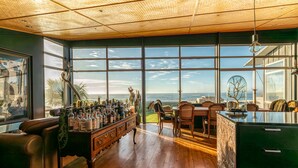 Una habitación amplia con ventanas grandes, piso de madera, una zona de bar con botellas y vistas al océano.