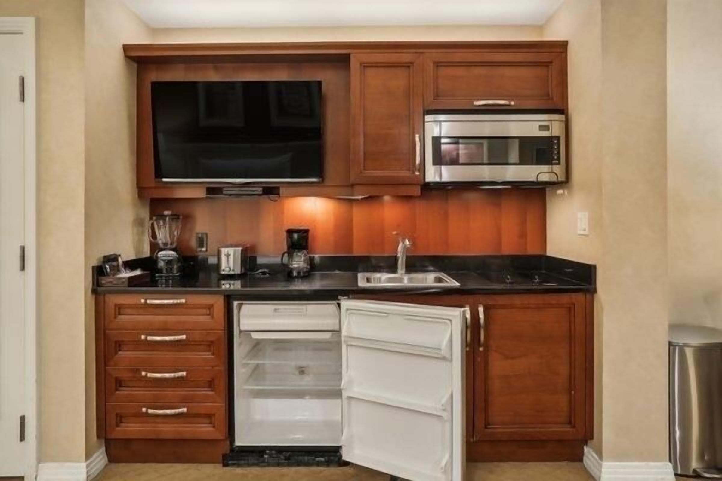 A compact kitchen with wooden cabinets, a built-in microwave, a refrigerator, and a flat-screen TV.