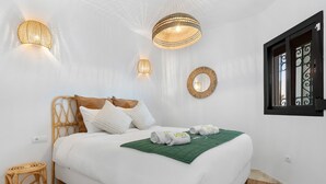 Une chambre à coucher bien aménagée, avec un lit, deux lampes fixées au mur, une fenêtre ornée de barreaux en fer décoratifs et une tête de lit en osier.