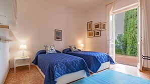 A bedroom with two beds, a small bedside table with a lamp, a window with a view of greenery, and a door leading to an outdoor area.
