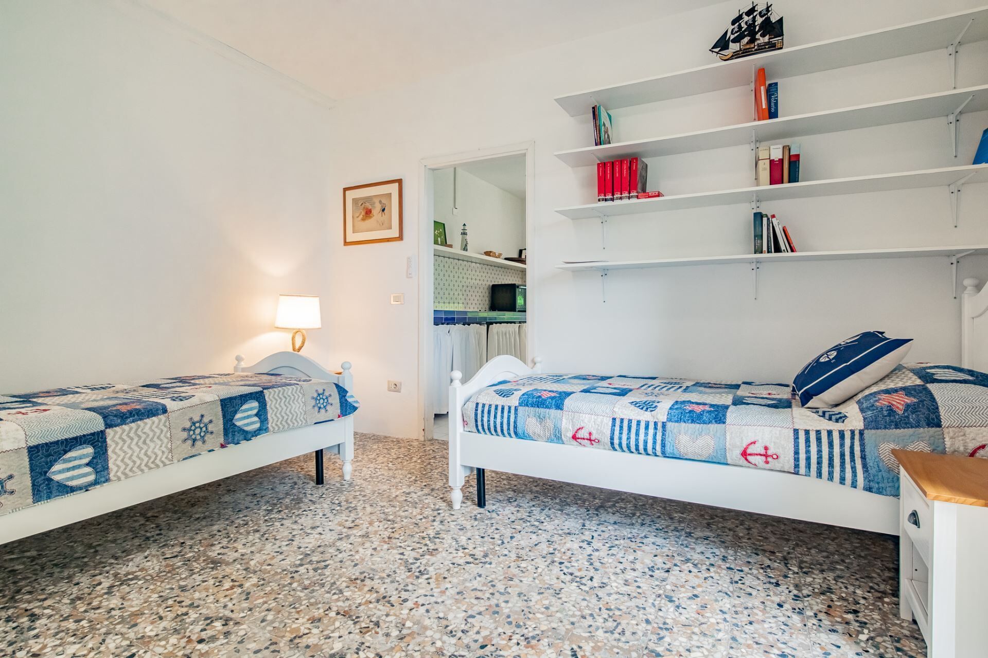 Un dormitorio con dos camas individuales, un armario blanco, una estantería y una cocina visible a través de una puerta abierta.